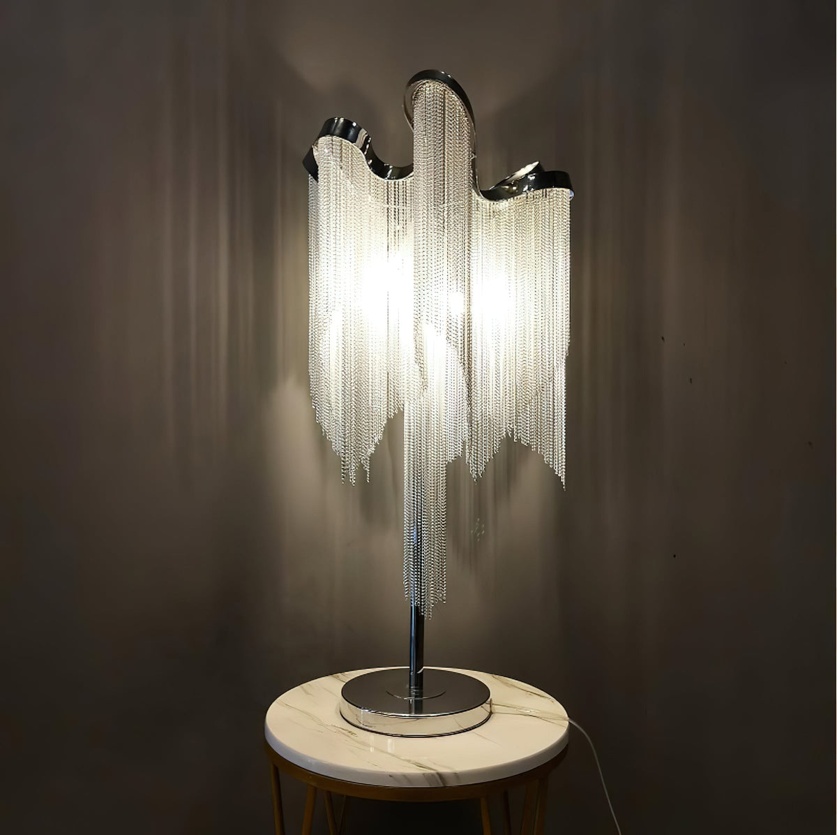 A lit Clérachic chain-fringe lamp on a round white marble-topped stand against a dark wall, its fringe glowing warm.