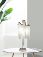 A Clérachic silver chain-fringe lamp on a round marble-top table with wooden legs, with a large green leaf at the window beside it.
