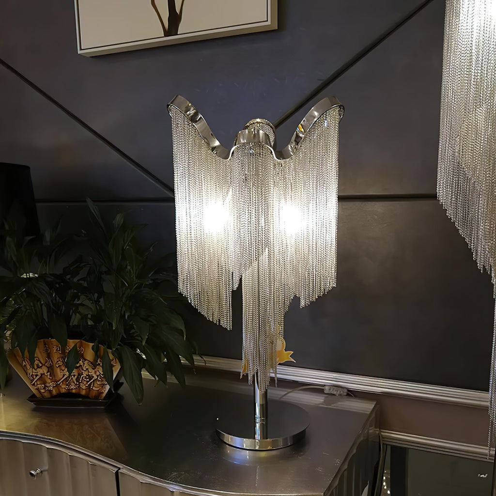Angled view of this Clérachic lamp lit on a dark dresser, with a green plant at left and chain fringe from a second light at the edge.