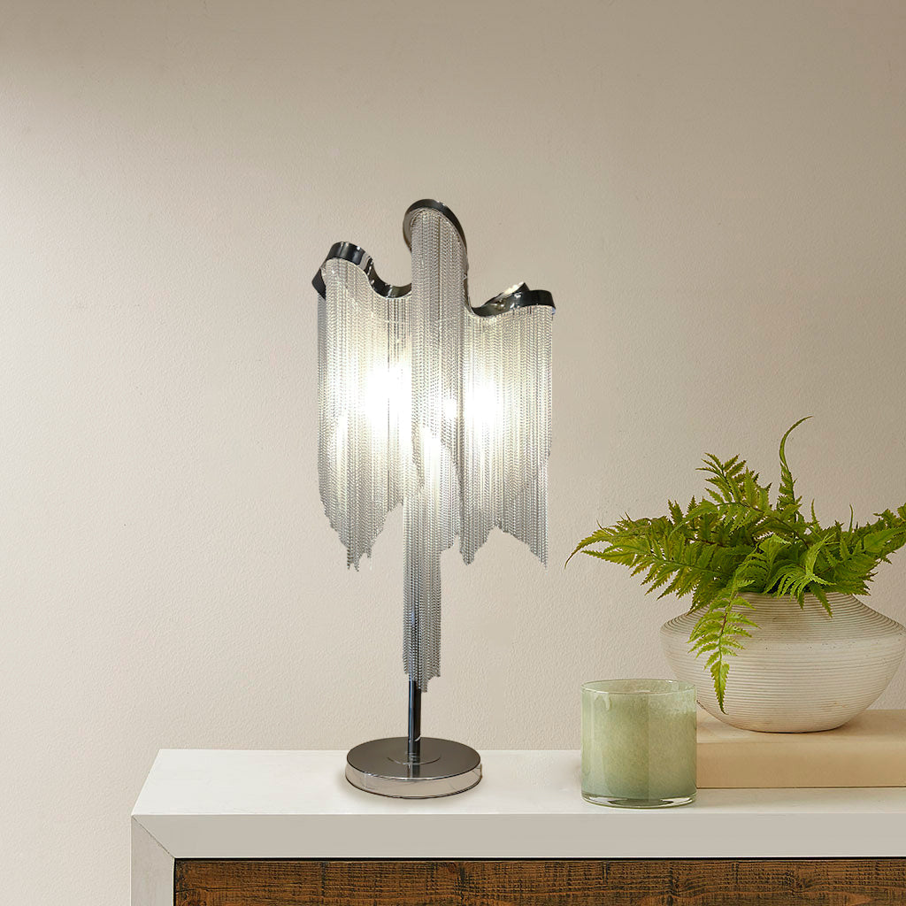 A Clérachic fringe lamp on a white shelf in warm light, with a green fern in a pot and a pale green candle next to it.