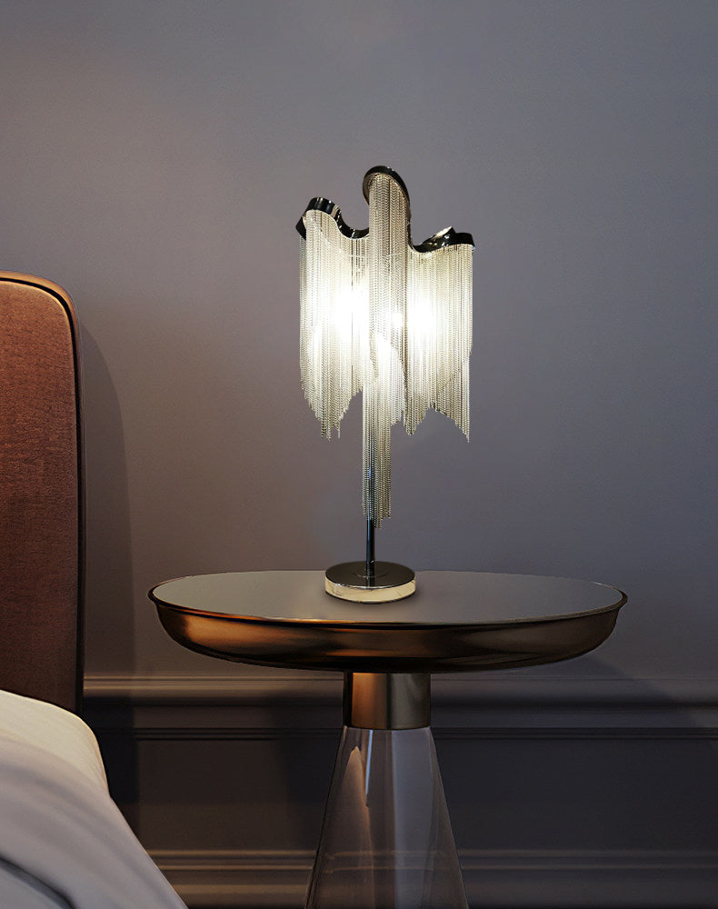 A Clérachic chain-fringe lamp on a round bronze-topped side table with a glass base, next to a rust-coloured headboard.