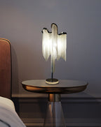 A Clérachic chain-fringe lamp on a round bronze-topped side table with a glass base, next to a rust-coloured headboard.