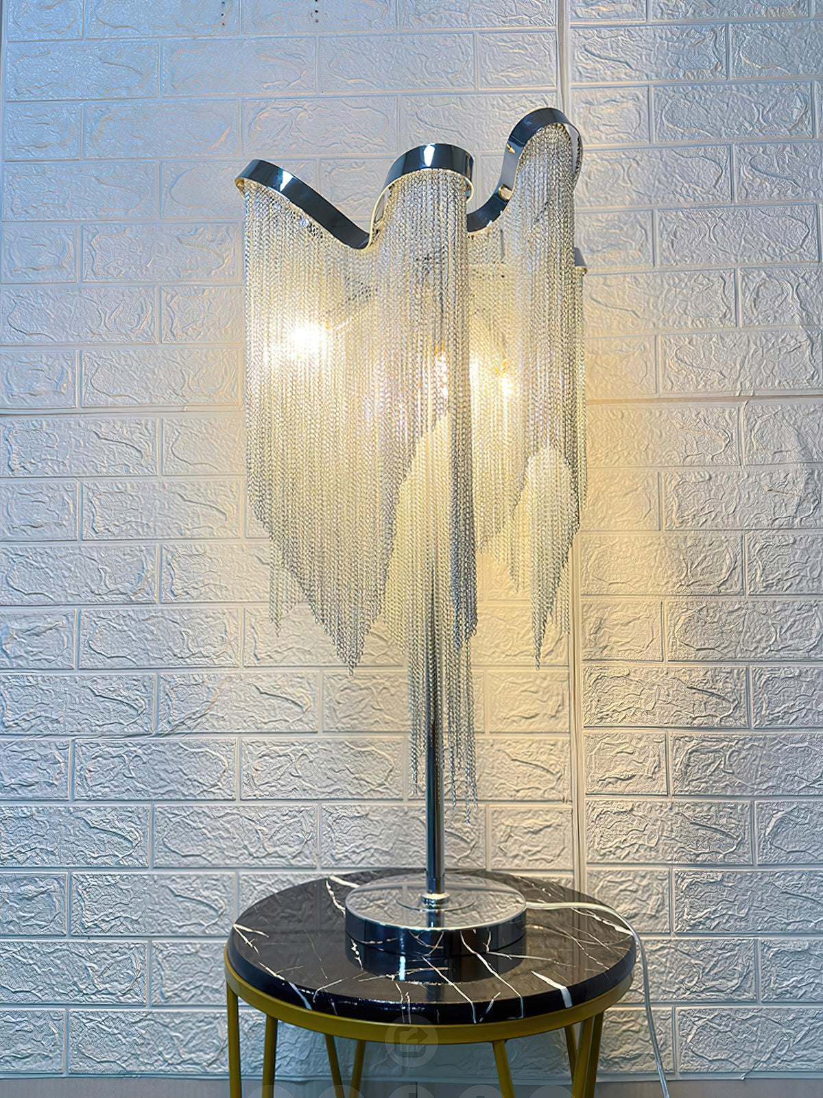 A lit Clérachic lamp on a black-marble stool with gold legs, in front of a white brick-pattern wall.