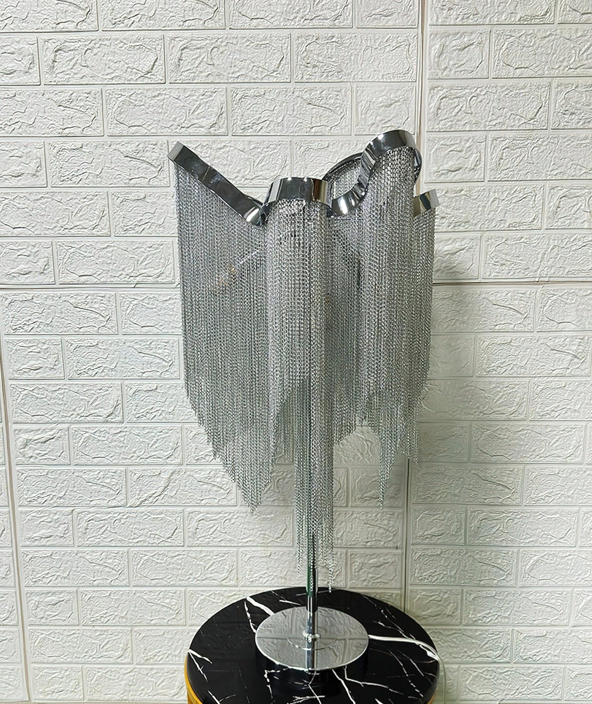 Rear three-quarter view of this Clérachic lamp unlit, its silver chains hanging in curtains over a black marble stool.