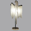 A Clérachic silver chain-fringe lamp lit against a mid-grey backdrop, showing the curved top, long chains and chrome base.