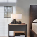 A lit Clérachic table lamp with a white shade and a dark mountain-shaped base, on a dark bedside table beside a bed and a framed artwork.