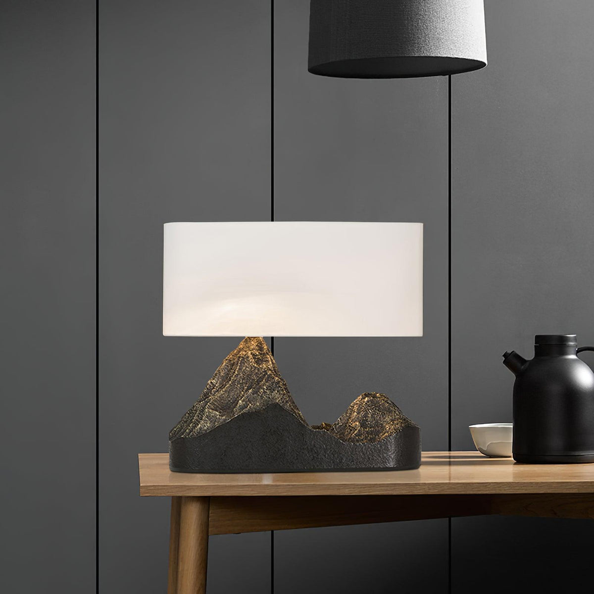 This Clérachic mountain-base table lamp glows under a white shade on a wooden side table, with a black jug and a bowl beside it.