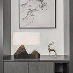 A Clérachic table lamp with a mountain-peak base on a grey cabinet, beside a brass deer figure and a large framed line drawing.
