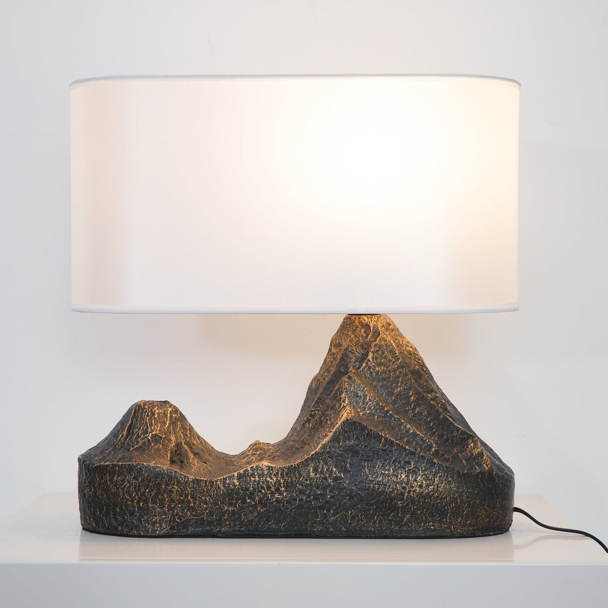 Close view of the lit white shade and the textured, bronze-flecked mountain base of this Clérachic table lamp on a white cabinet.
