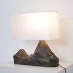A Clérachic table lamp lit warm, its twin-peak resin base showing bronze highlights, with the cord trailing off a white cabinet.