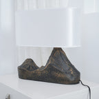 Low side view of this Clérachic table lamp, its broad white shade above a ridged twin-peak base, with a black cord on a white surface.