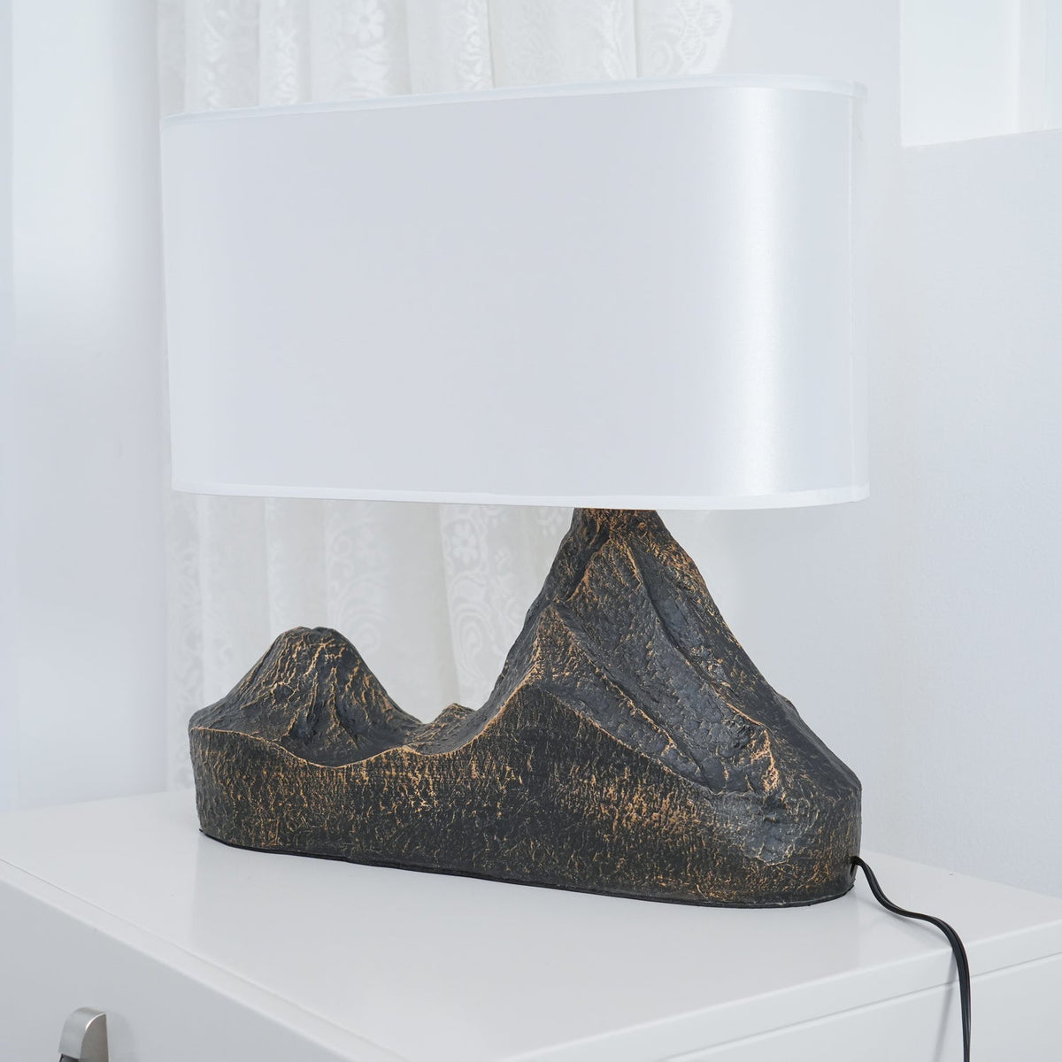Low side view of this Clérachic table lamp, its broad white shade above a ridged twin-peak base, with a black cord on a white surface.