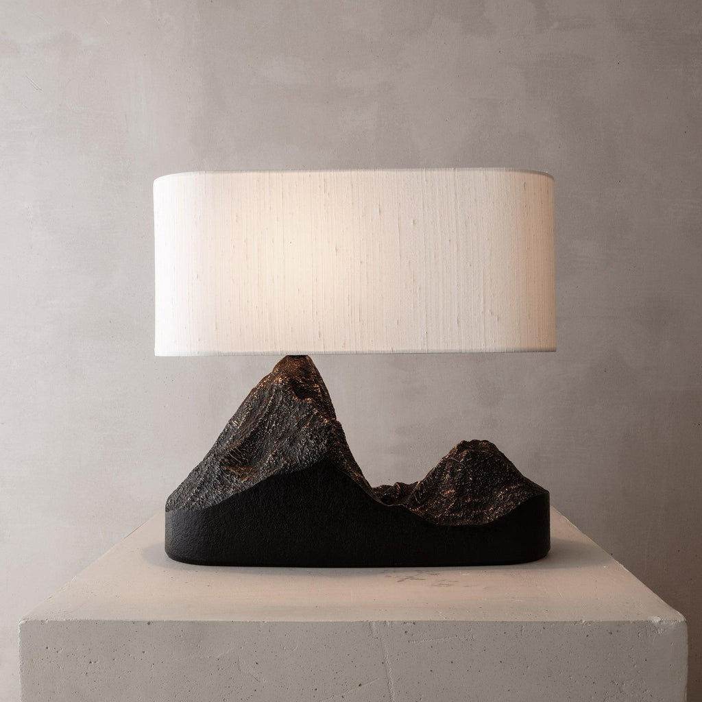 A Clérachic table lamp with a white woven-look shade and a black mountain base, on a pale stone block against a grey wall.