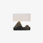 A small, straight-on Clérachic table lamp, a pale rectangular shade above a black base with two bronze-toned peaks, on white.