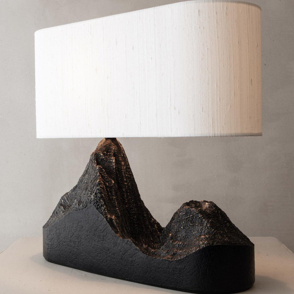 Three-quarter view of the cream shade and the carved peaks on the black base of this Clérachic table lamp, against a grey wall.