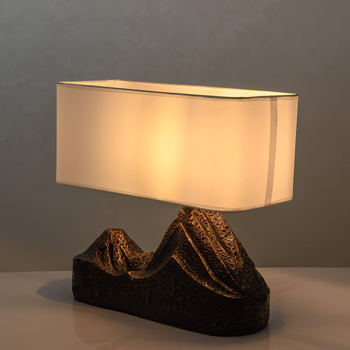 The shade of this Clérachic table lamp glows warm and golden above a dark, ridged twin-peak base, on a pale ledge.