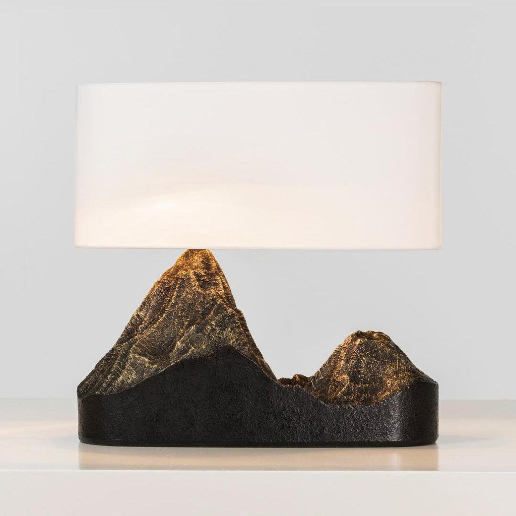 A Clérachic table lamp with a wide white shade and a dark base whose two peaks glow bronze, on a pale surface.