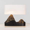 A Clérachic table lamp with a wide white shade and a dark base whose two peaks glow bronze, on a pale surface.