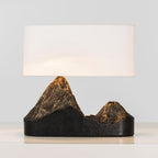 A Clérachic table lamp with a wide white shade and a dark base whose two peaks glow bronze, on a pale surface.