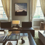 A lit Clérachic table lamp sits low on a dark coffee table of books, between leather armchairs in a bright room with tall windows.