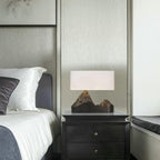 A Clérachic table lamp with a white shade and a dark mountain base on a black bedside chest, beside a bed with white pillows.