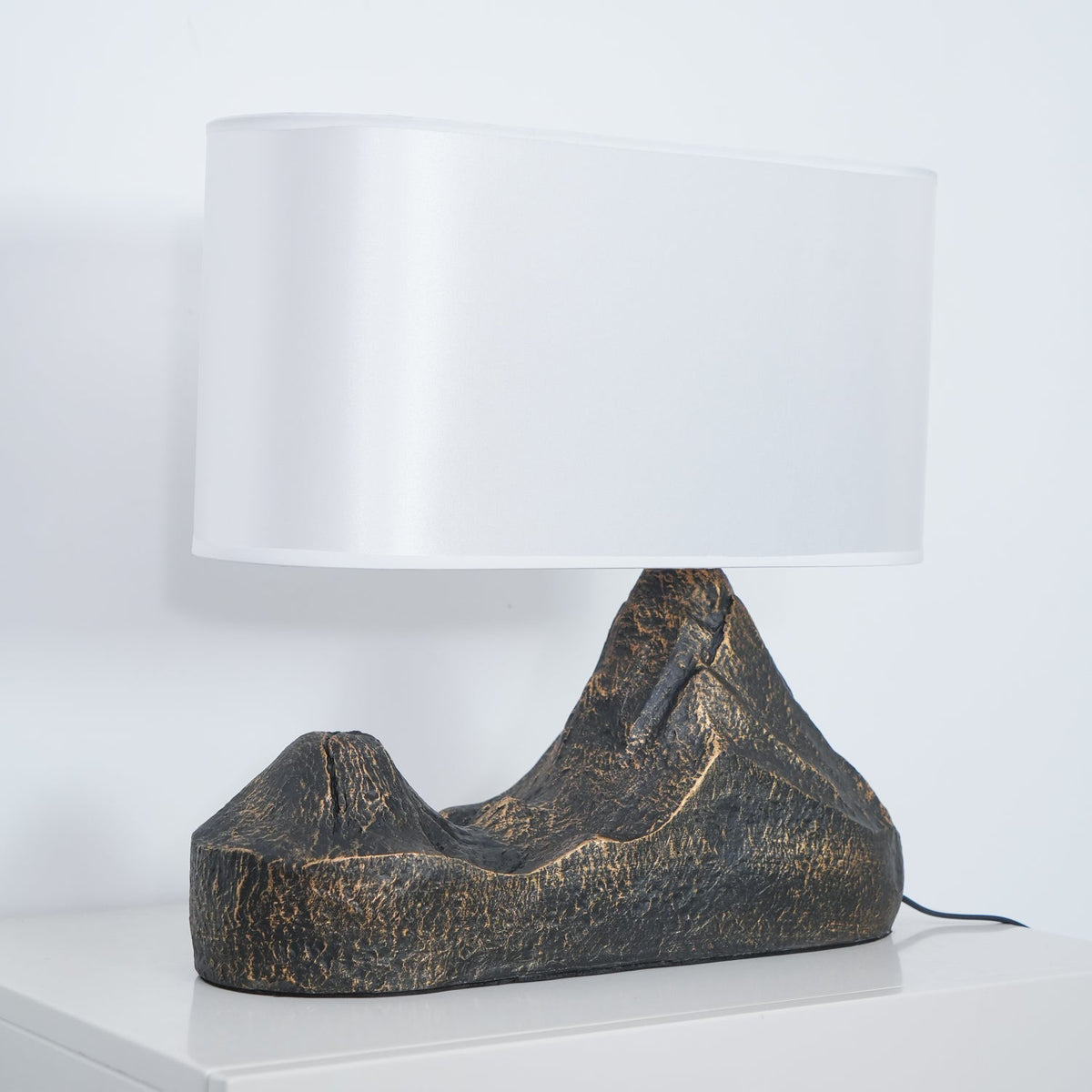 Side view of a Clérachic table lamp, a broad white shade above a ridged bronze-brown base, with its black cord on a white cabinet.