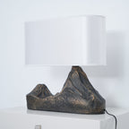 A Clérachic table lamp straight on, with a white shade and a rugged twin-peak base in dark bronze-brown, on a white cabinet.
