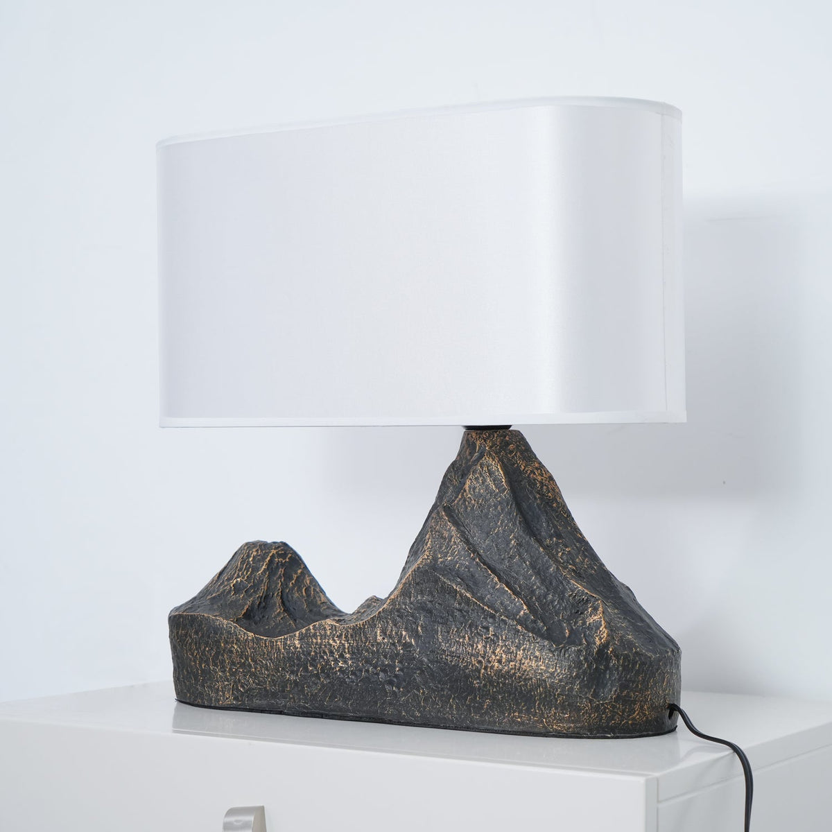 A Clérachic table lamp straight on, with a white shade and a rugged twin-peak base in dark bronze-brown, on a white cabinet.