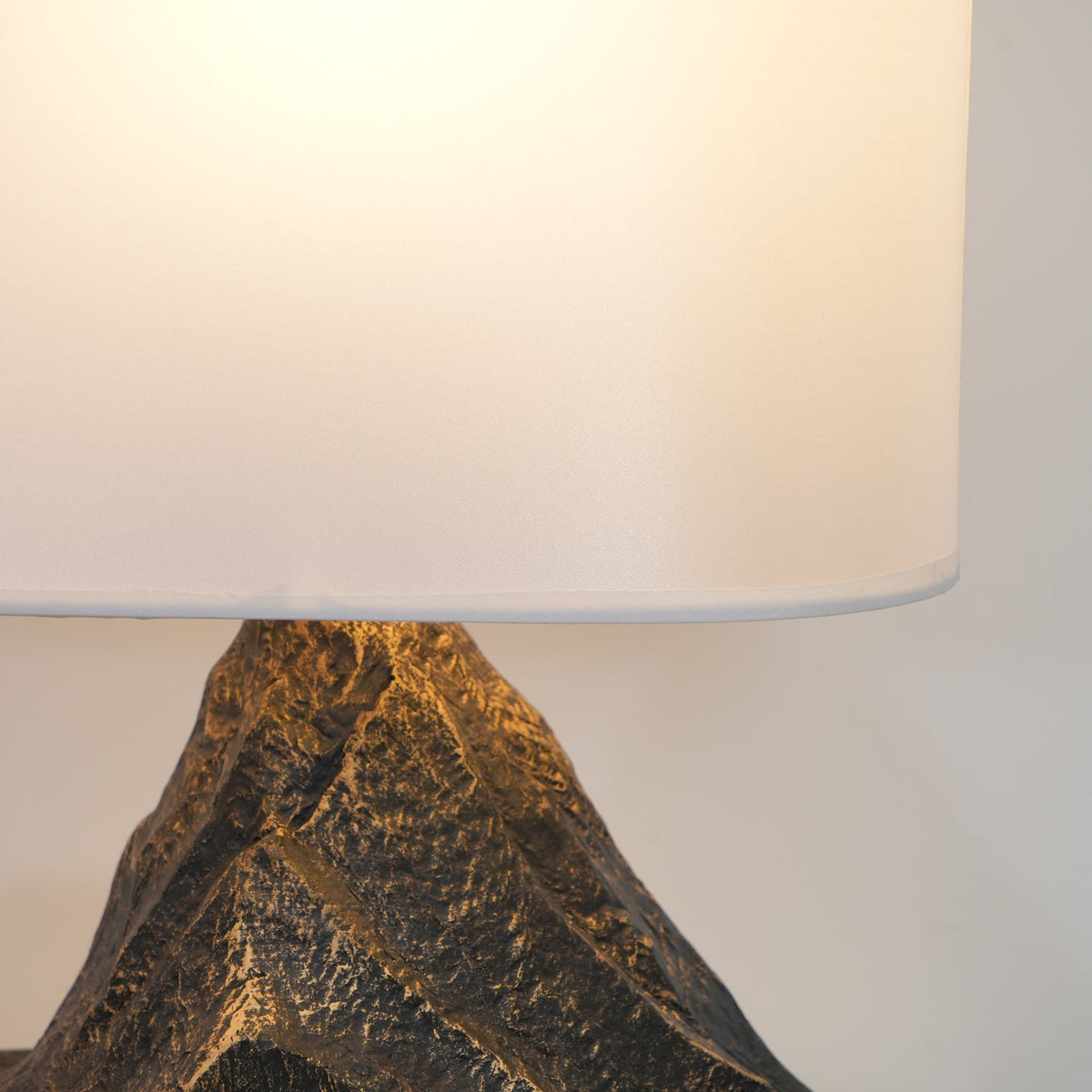 Close view of the lit white shade and the top of the carved bronze-brown peak on this Clérachic table lamp.