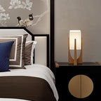 Clérachic table lamp in natural wood on a black bedside cabinet, its rectangular shade lit beside a bed.