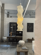 Clérachic crushed-paper wave-column pendant light hanging the full height of a plaster-walled loft, above a dark glazed cabinet, a black canvas and stoneware pots.