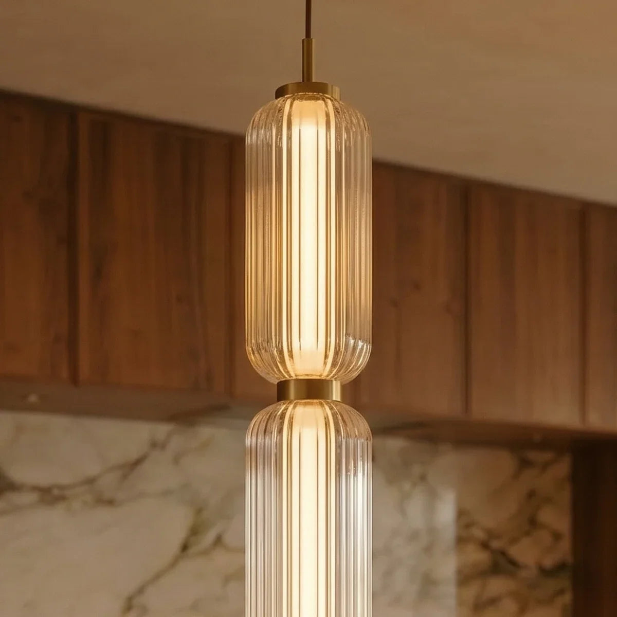 Close up of Clérachic ribbed clear glass pendant tiers with a brass-gold collar, lit warmly in a timber kitchen room.