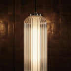 Close up of a lit Clérachic ribbed clear glass pendant with a chrome cap and black cable against dark tiled walls.