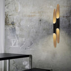 Two long gold-and-black tubes of this Clérachic wall light rise on a weathered grey concrete wall above a dark table edge.