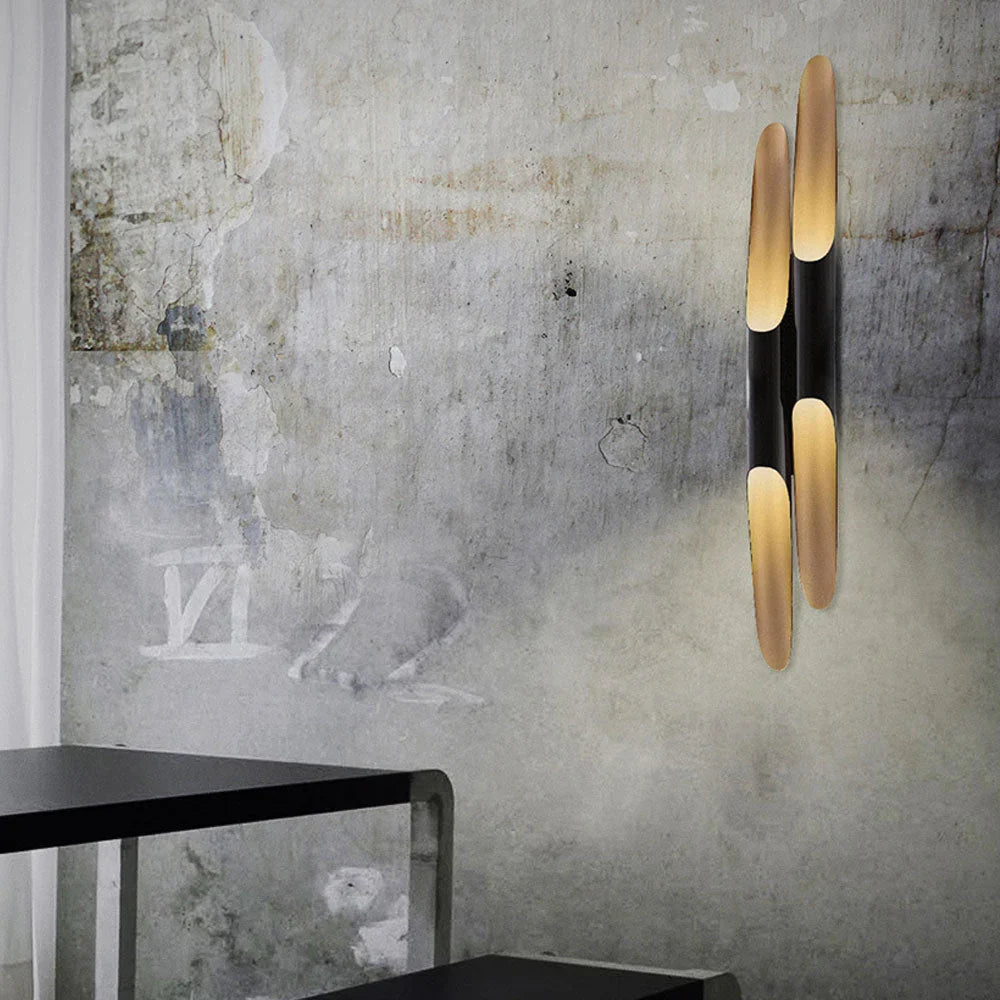 Two long gold-and-black tubes of this Clérachic wall light rise on a weathered grey concrete wall above a dark table edge.