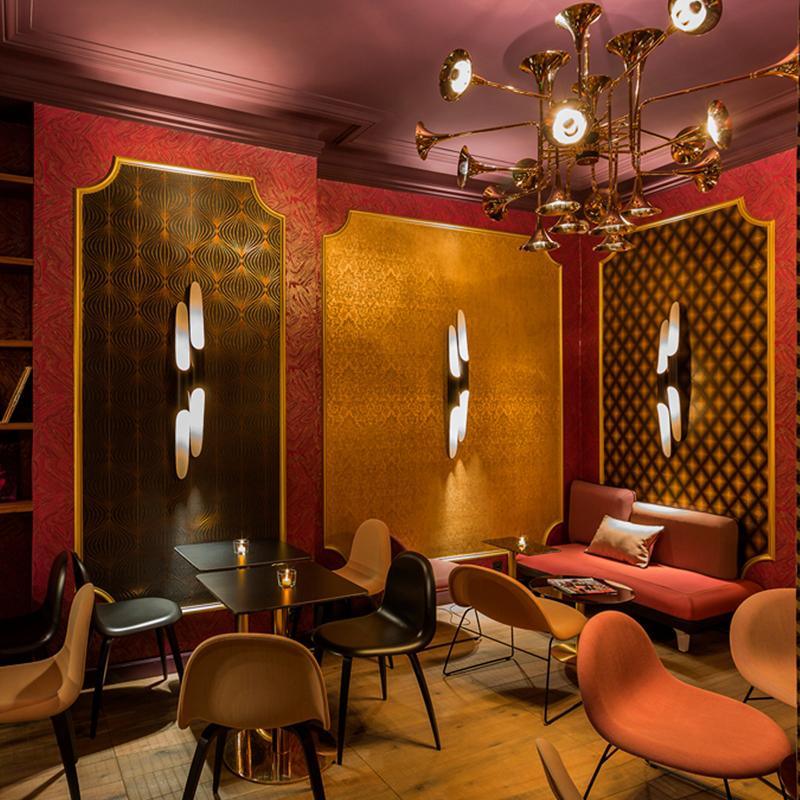 In a red and gold lounge with rust-coloured chairs, three Clérachic tube wall lights glow white against patterned wall panels.