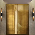 A Clérachic tube wall light stands at each side of a pair of brushed brass doors, with warm gold light from each open mouth.