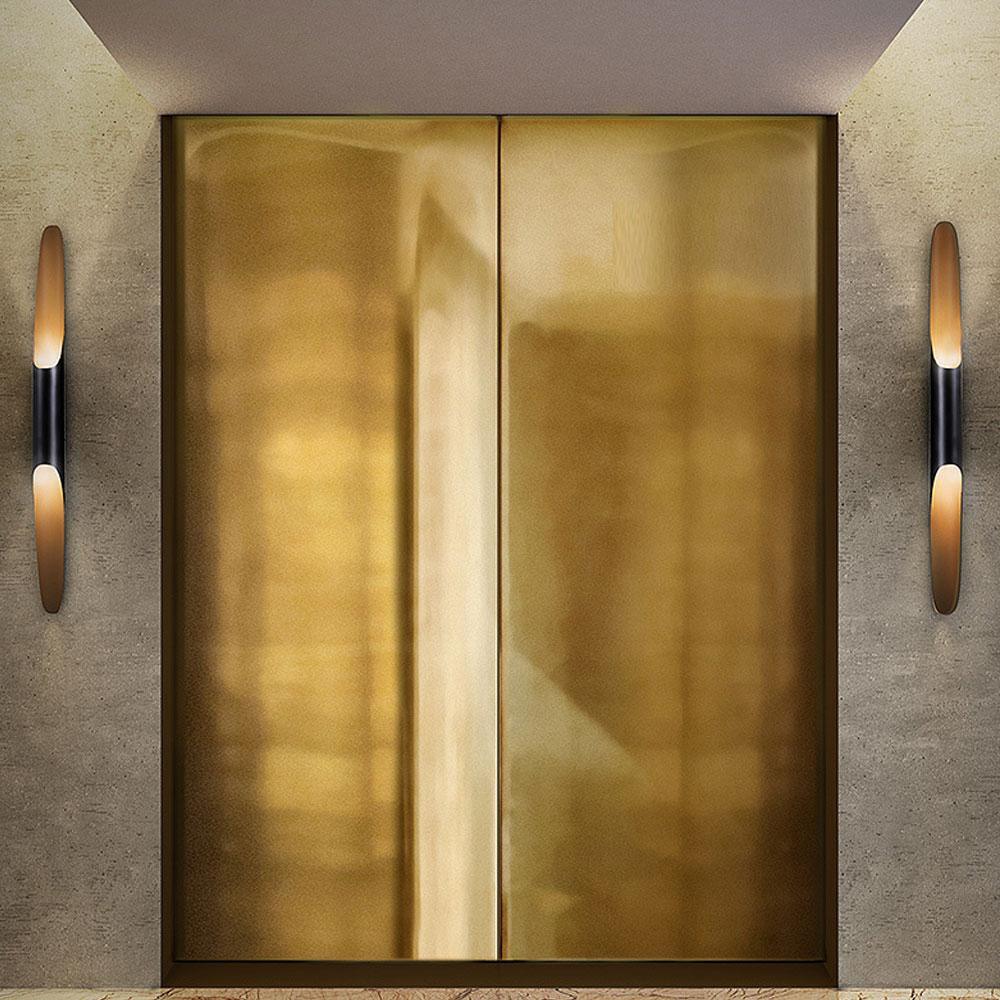 A Clérachic tube wall light stands at each side of a pair of brushed brass doors, with warm gold light from each open mouth.