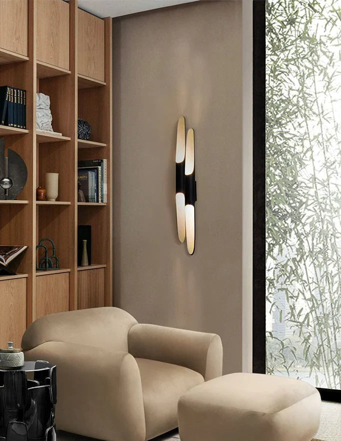 Next to wooden shelves and a cream armchair, a Clérachic double-tube wall light glows against a warm taupe wall and a bamboo view.