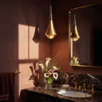 Clérachic gold tapered pendant light hung beside a gilt mirror, with calla lilies and a dark marble vanity top.