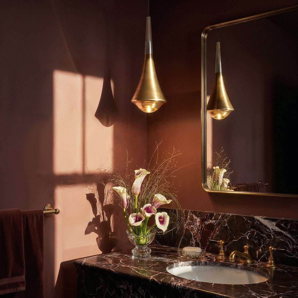 Clérachic gold tapered pendant light hung beside a gilt mirror, with calla lilies and a dark marble vanity top.