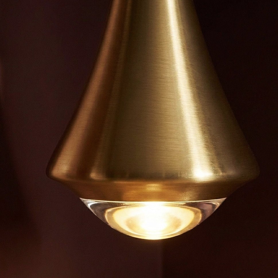 Close view of the brushed gold Clérachic pendant, its softly rounded clear glass base glowing warm against a dark wall.