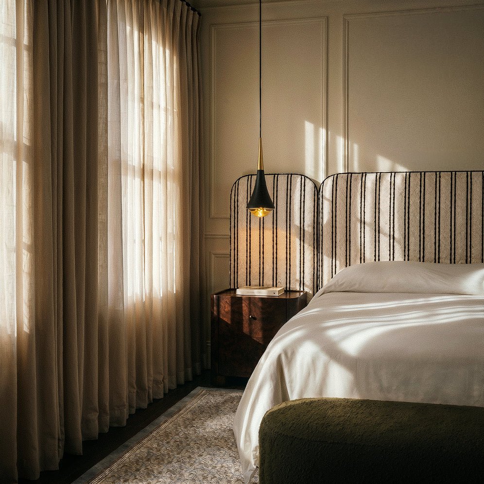 Clérachic black and gold tapered pendant hung by a walnut bedside table, striped headboard and sheer linen curtains.
