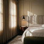 Clérachic black and gold tapered pendant hung by a walnut bedside table, striped headboard and sheer linen curtains.