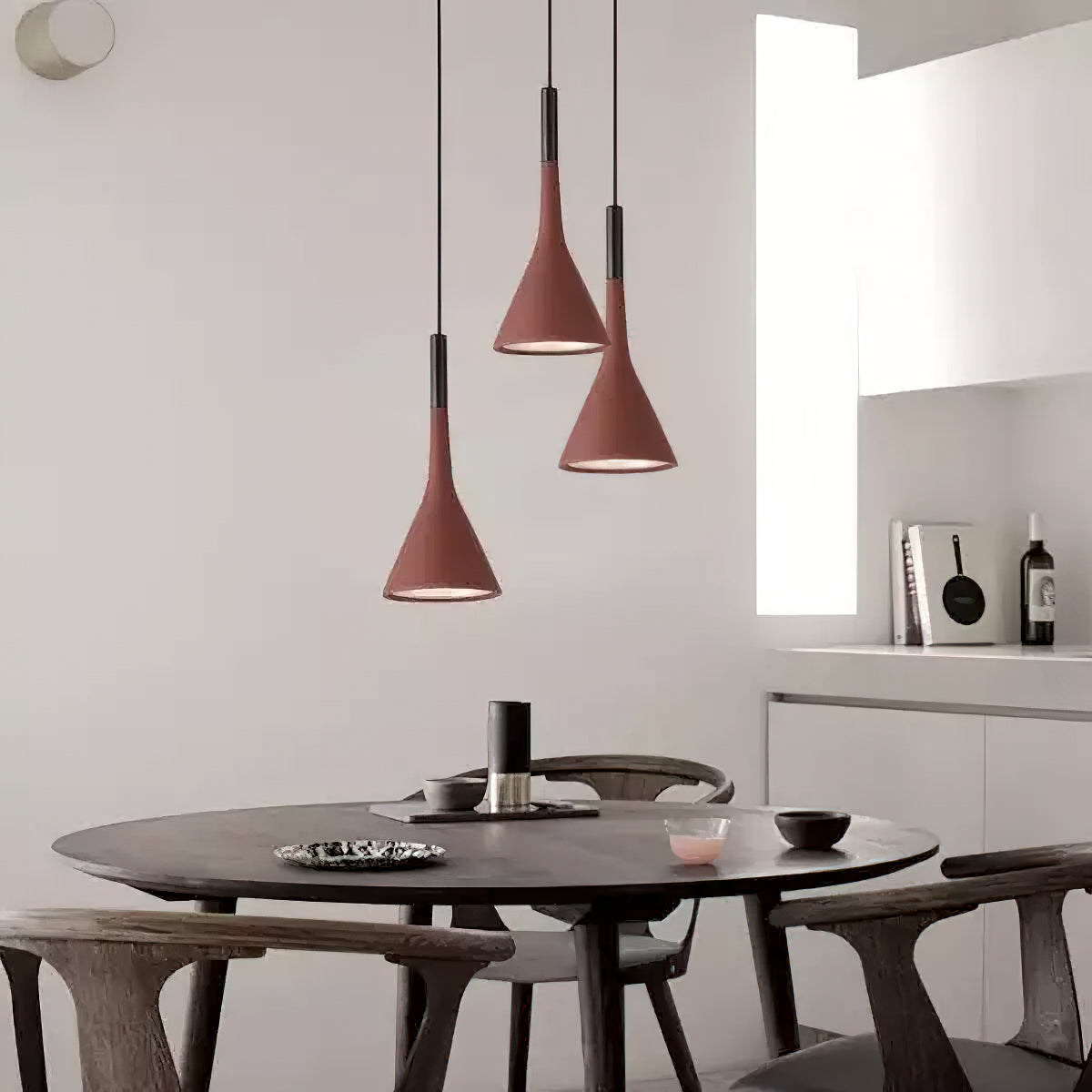 Three brick-red concrete Clérachic pendants at different heights over a round dark dining table, with a white kitchen behind.