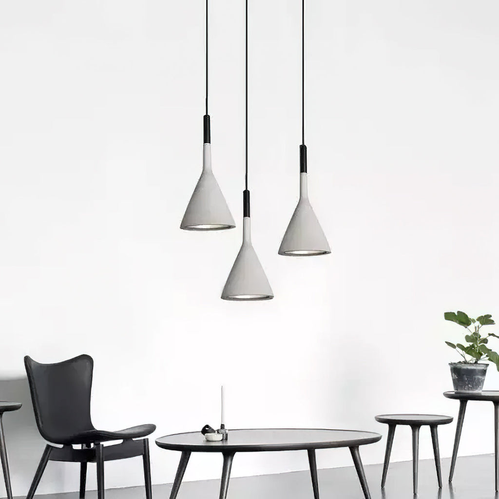 Three grey concrete Clérachic pendants hung at staggered heights above a low black table and slim-legged chairs against a white wall.
