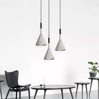Three grey concrete Clérachic pendants hung at staggered heights above a low black table and slim-legged chairs against a white wall.