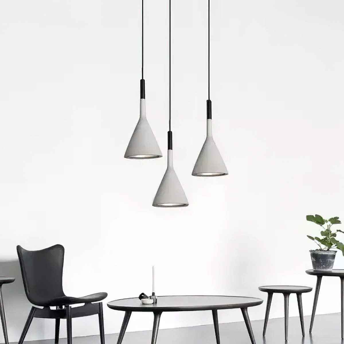 Three grey concrete Clérachic pendants hung at staggered heights above a low black table and slim-legged chairs against a white wall.
