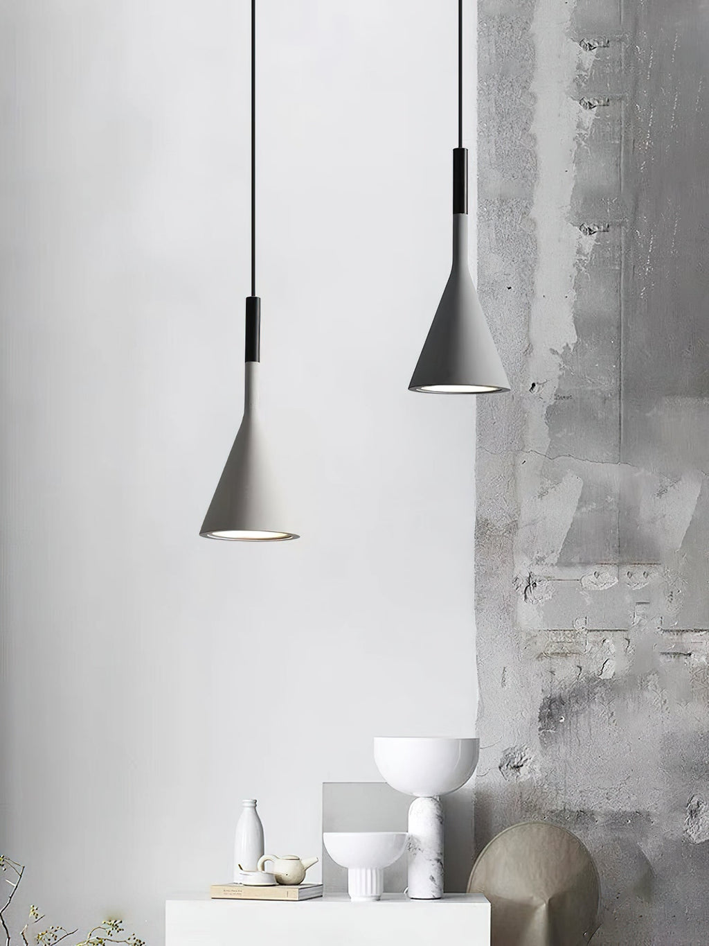 Two grey concrete Clérachic pendants beside a rough grey plaster wall, above a white shelf with sculptural bowls and vases.