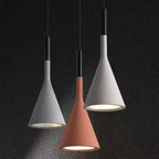 Three Clérachic concrete pendants in two greys and a terracotta orange against a dark textured wall, each lit with a silver-lined opening.
